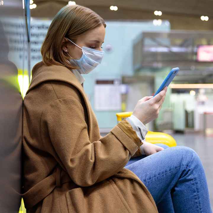 A woman wearing a face mask sits at an airport while scrolling through her phone.